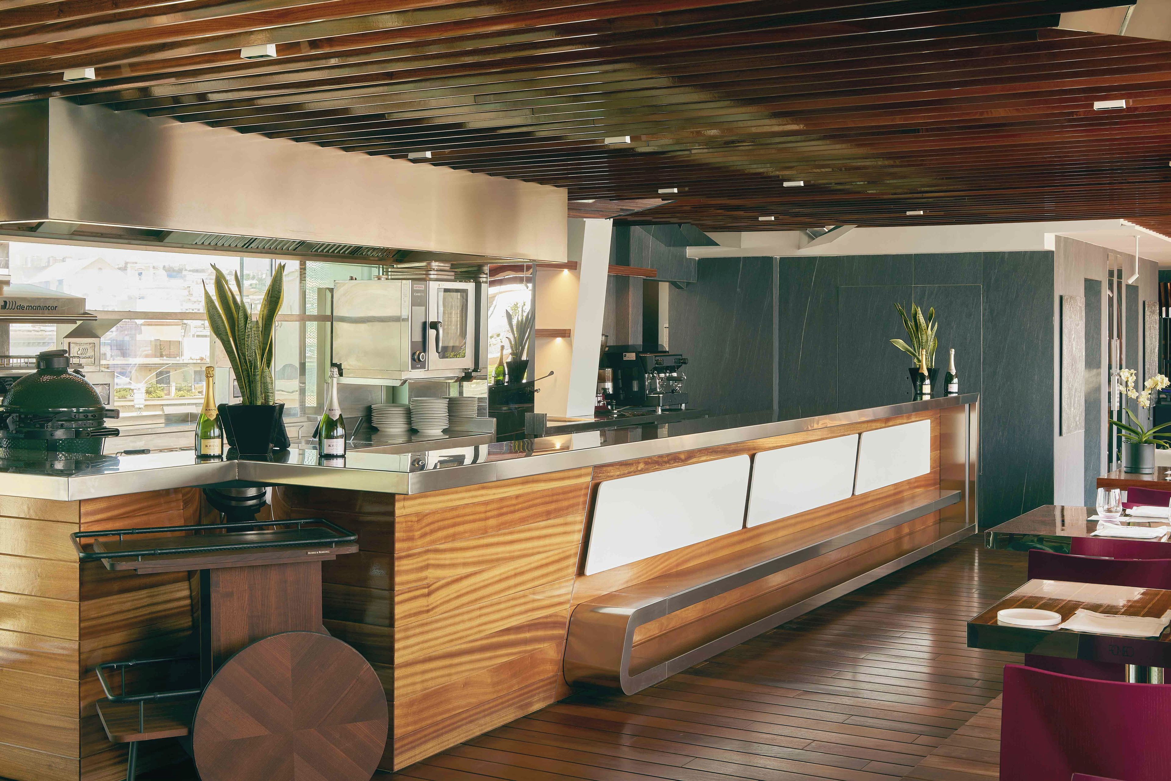 Interior moderno de restaurante con mostrador de madera, zona de estar y vistas a la ciudad a través de amplios ventanales.