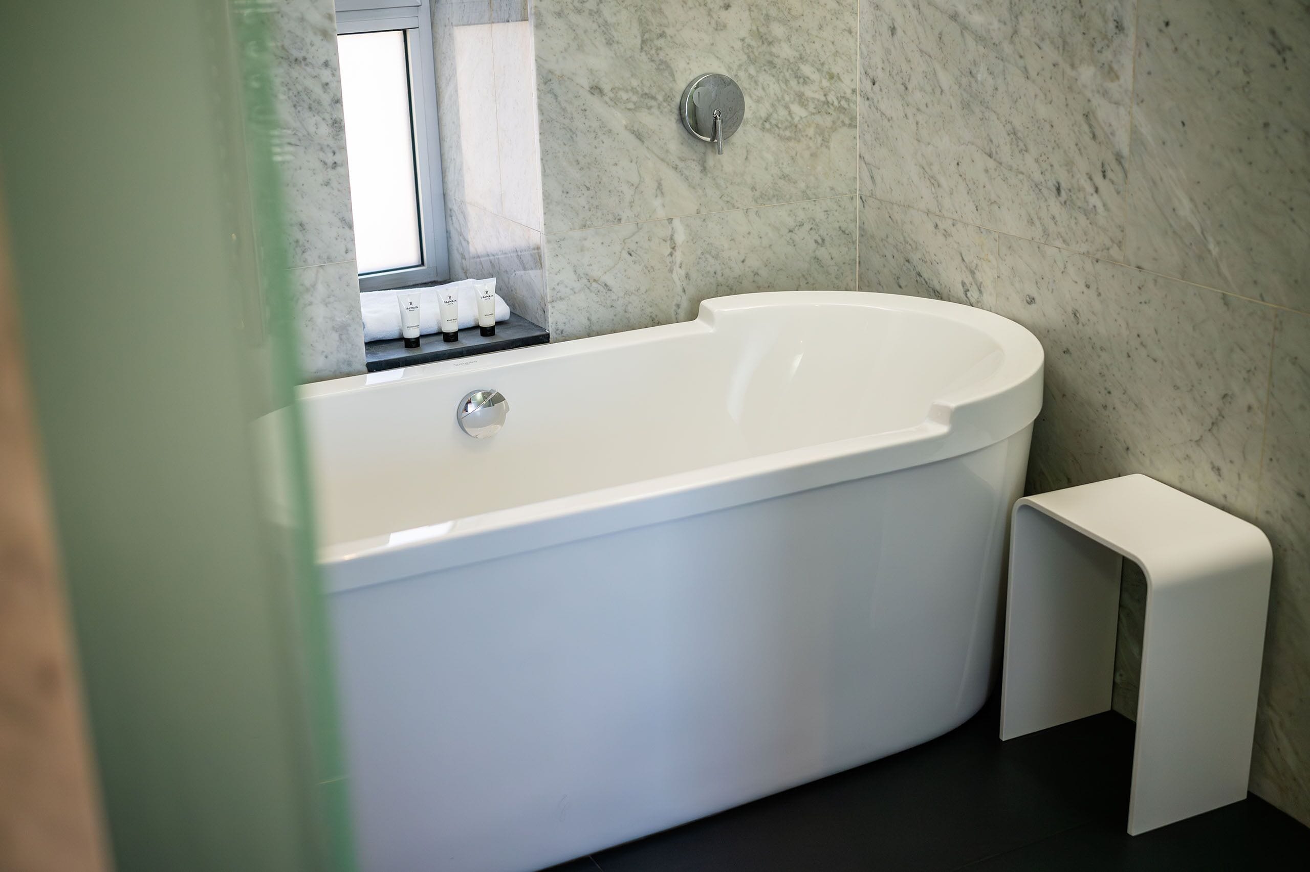 A modern bathroom with a freestanding bathtub, a small stool, and a window with a view.