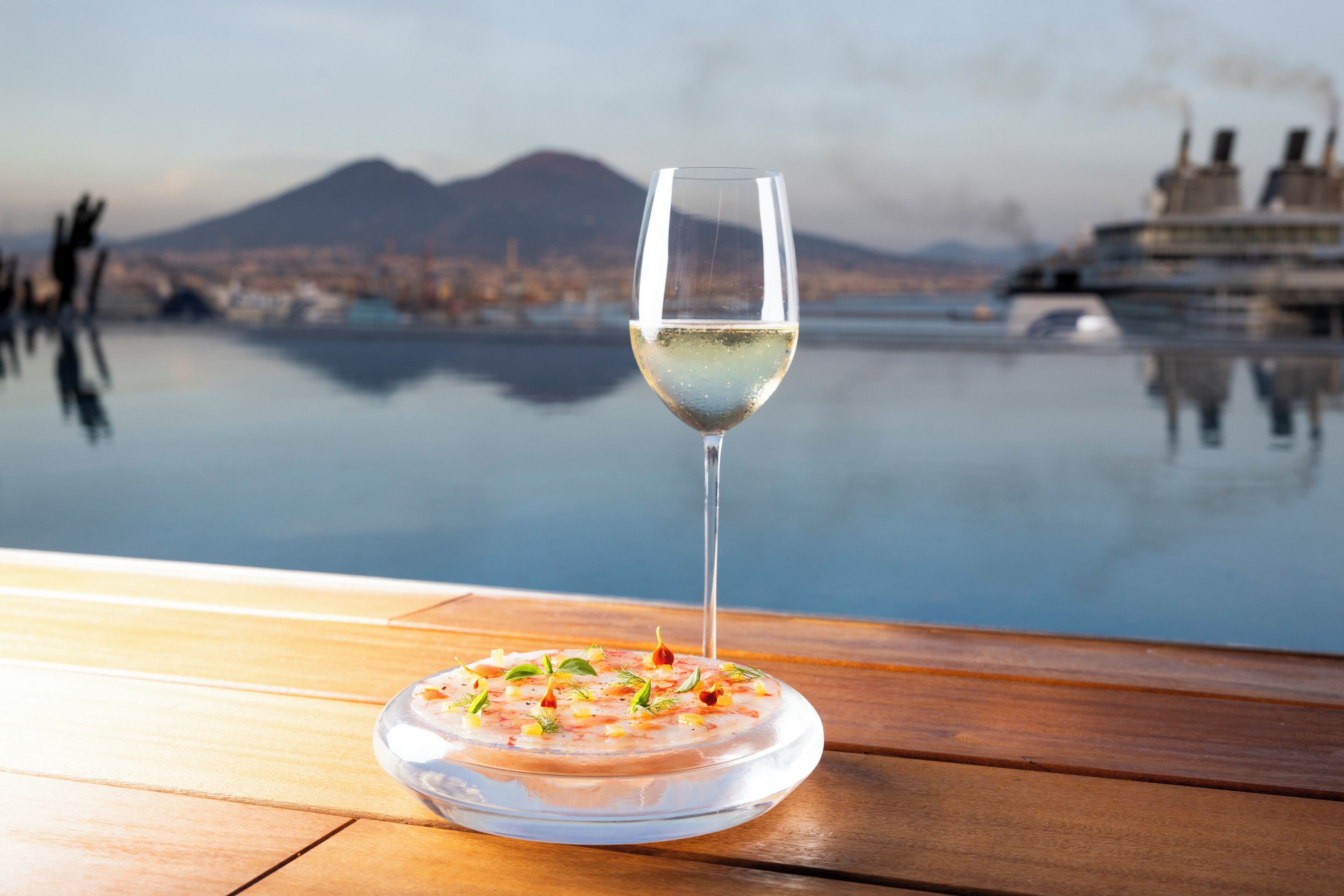 A clear glass of white wine on a wooden table with a view of a calm body of water and a cruise ship in the background.