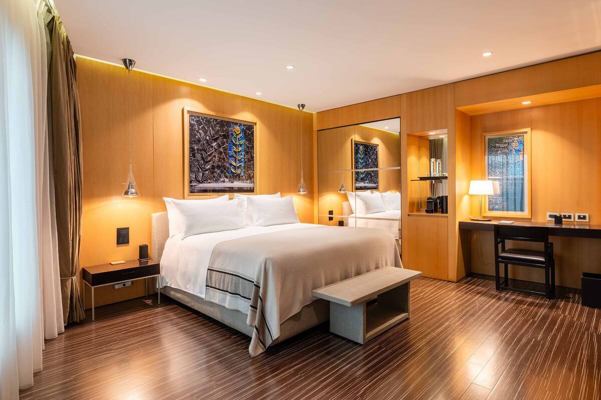 A hotel room with a large bed, bedside tables, a desk, and a bench.