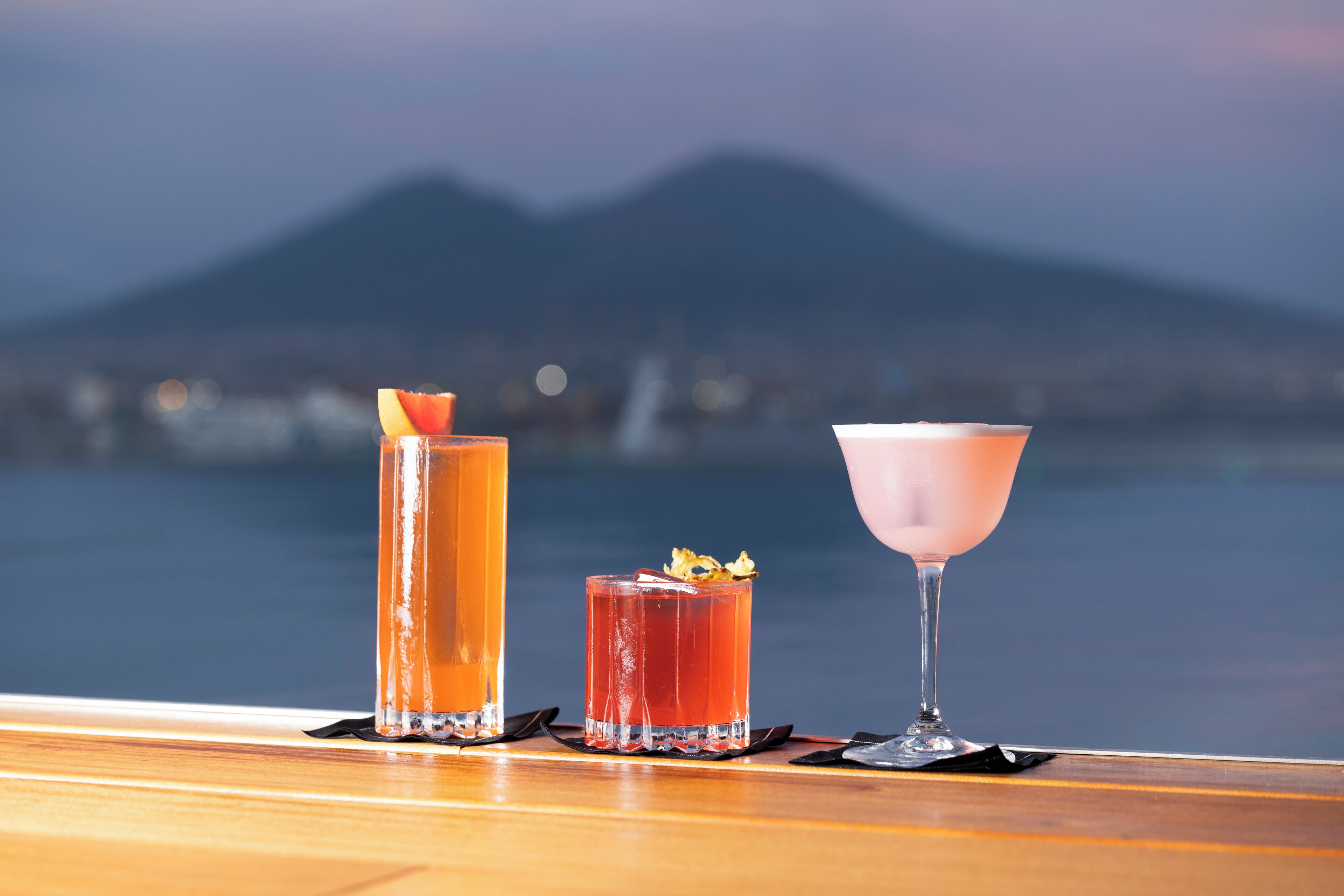 Three cocktails on a wooden bar counter with a view of a mountain and water in the background.