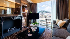 A modern hotel room with a fireplace, a flat-screen TV, a sofa, and a large window offering a city view.