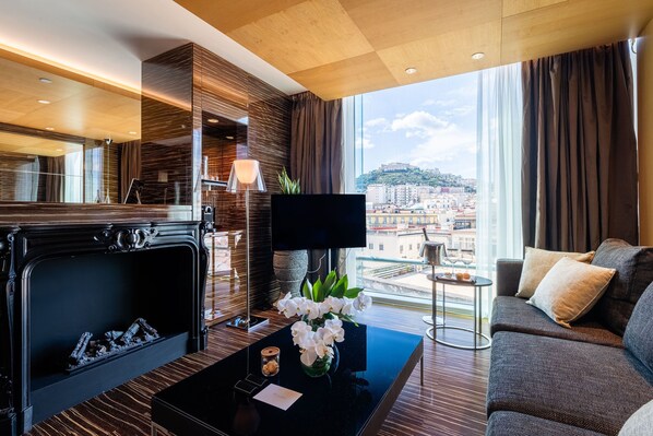 A modern hotel room with a fireplace, a flat-screen TV, a sofa, and a large window offering a city view.