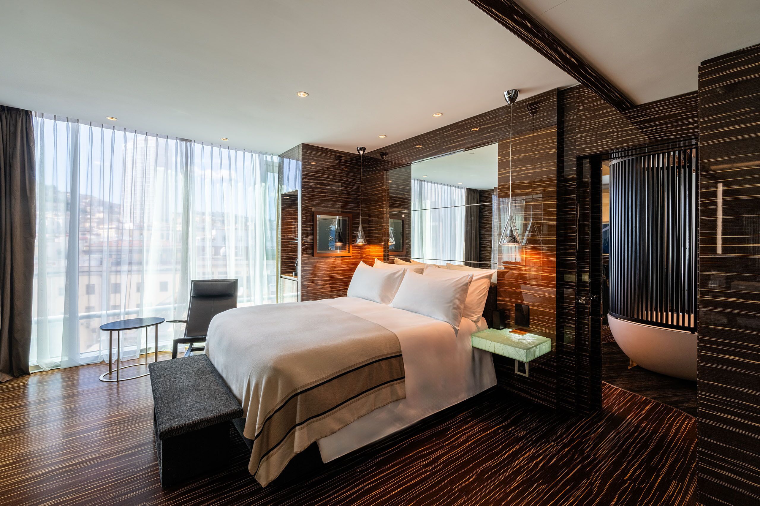A modern hotel room with a large bed, a small table, a bench, a bathtub, and a large window with curtains.