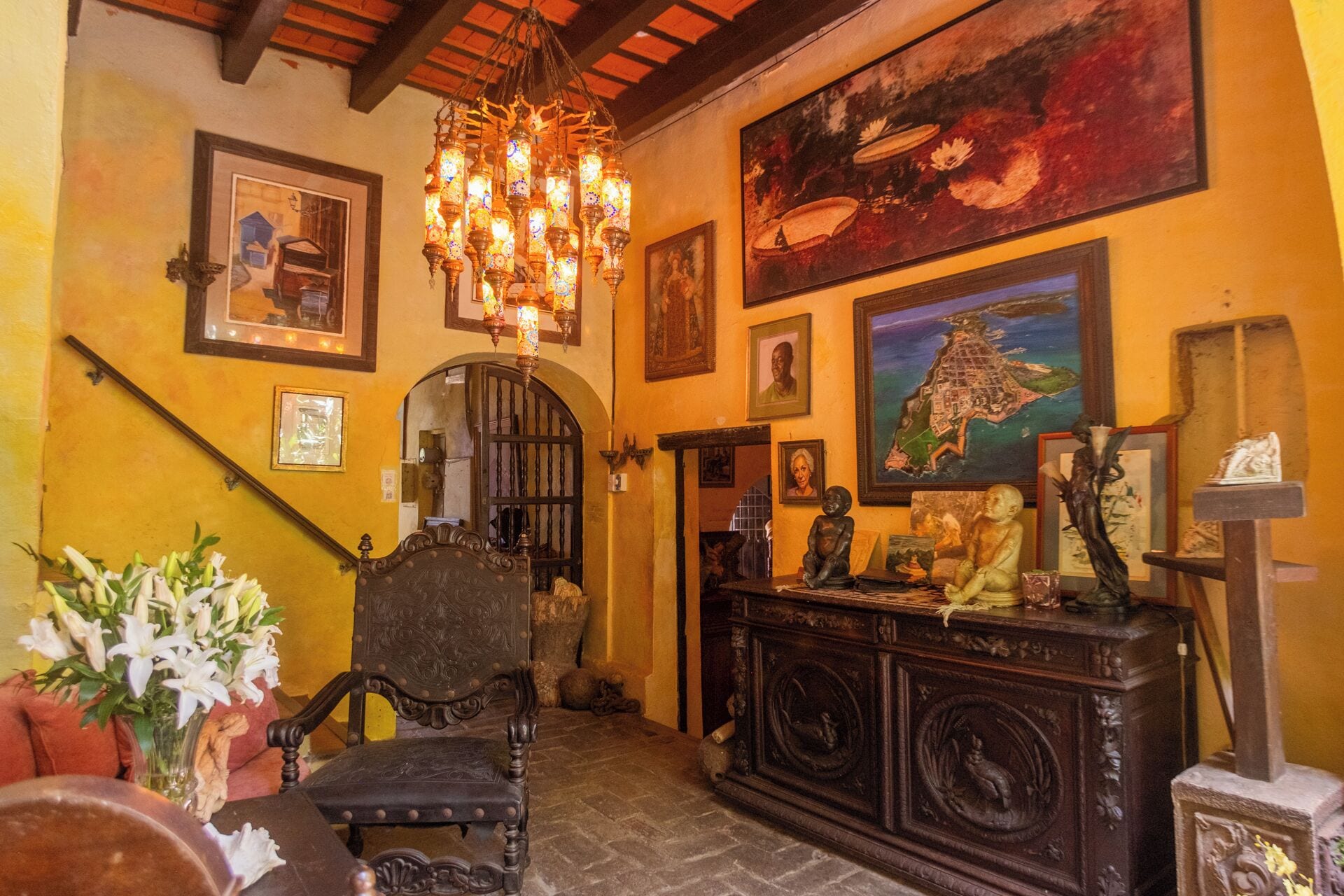 Una habitación con una escalera, una araña, una silla de madera, un armario de madera y varias fotografías enmarcadas en las paredes.