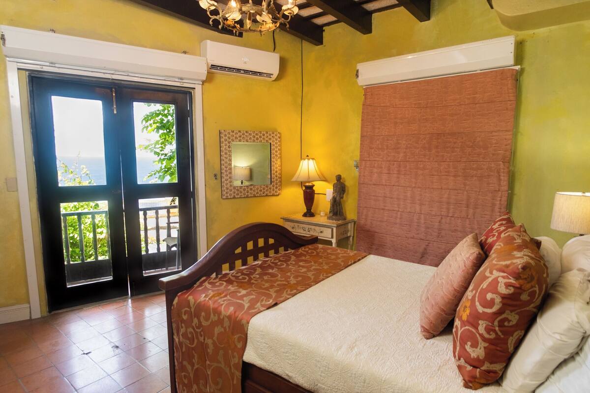 A room with a bed, a nightstand, a lamp, a mirror, and a window with a view of greenery.