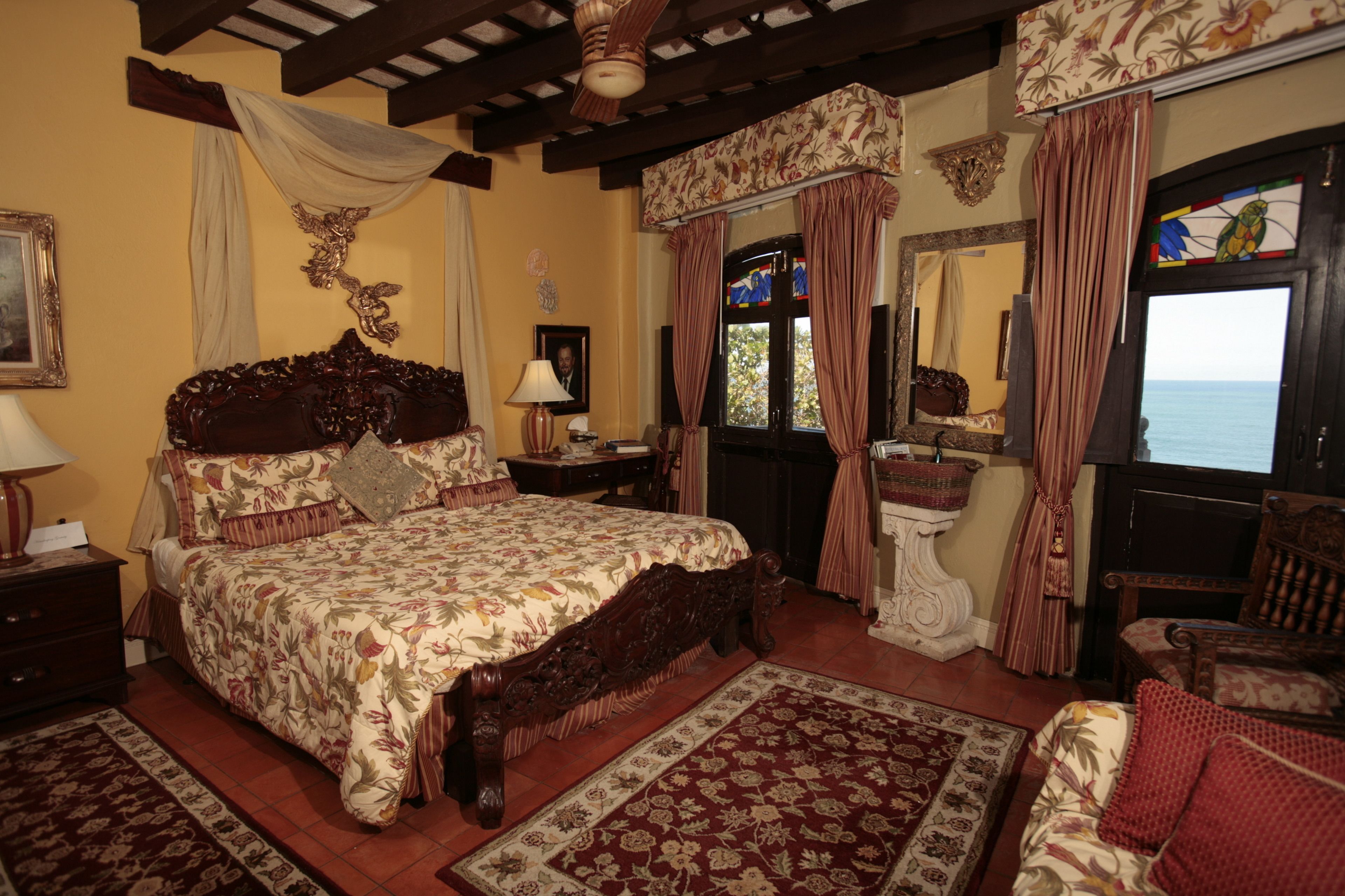 Un dormitorio tradicional con una cama grande, cabecera de madera, mesilla de noche, espejo, ventana con cortinas y vista al mar.