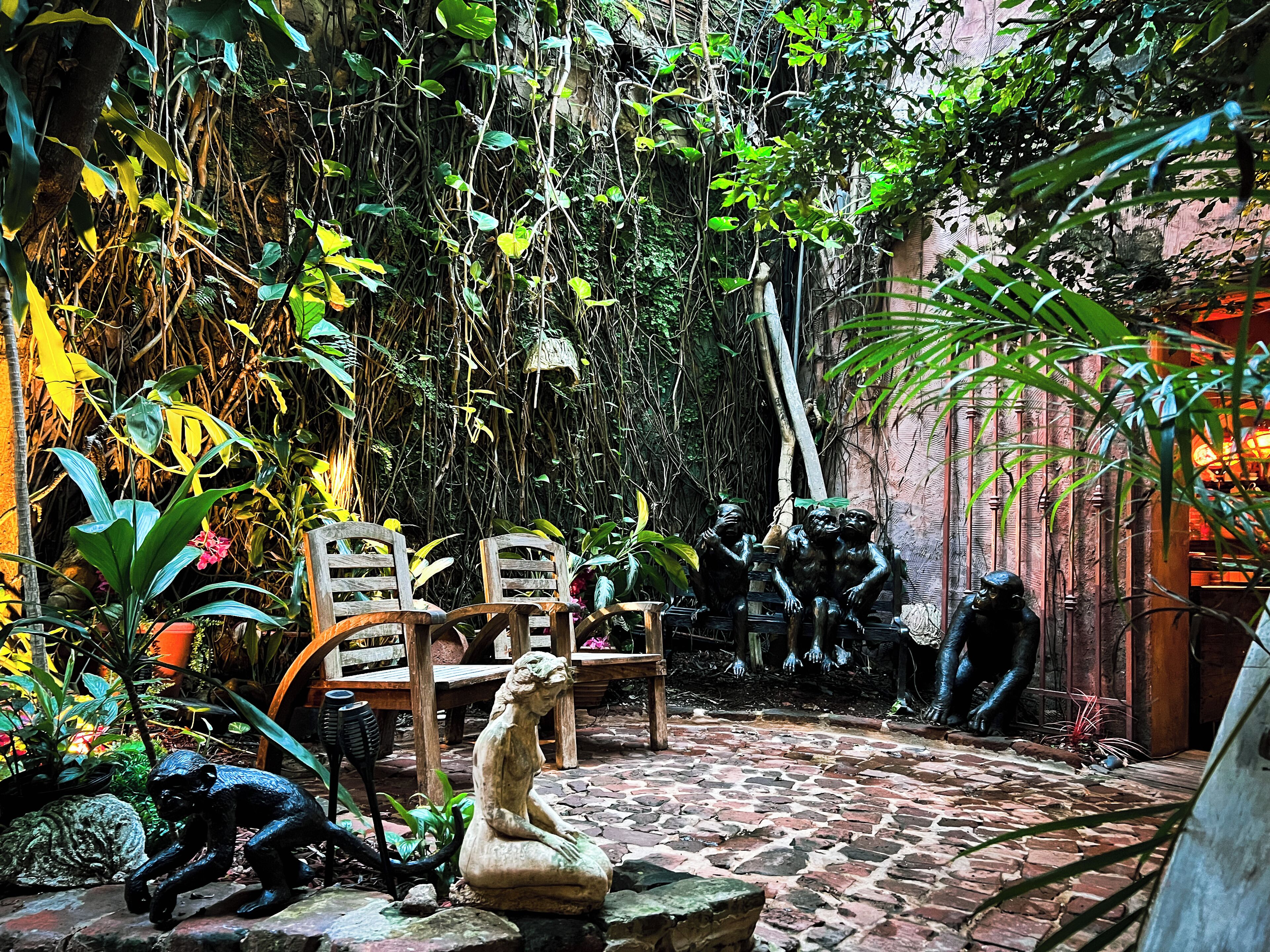 A courtyard with a stone floor, wooden chairs, and a statue of a seated figure.