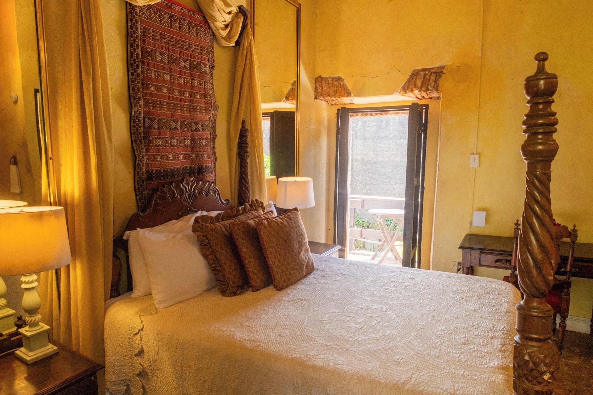 A bedroom with a large bed, a wooden headboard, a patterned curtain, and a view of the outside balcony.