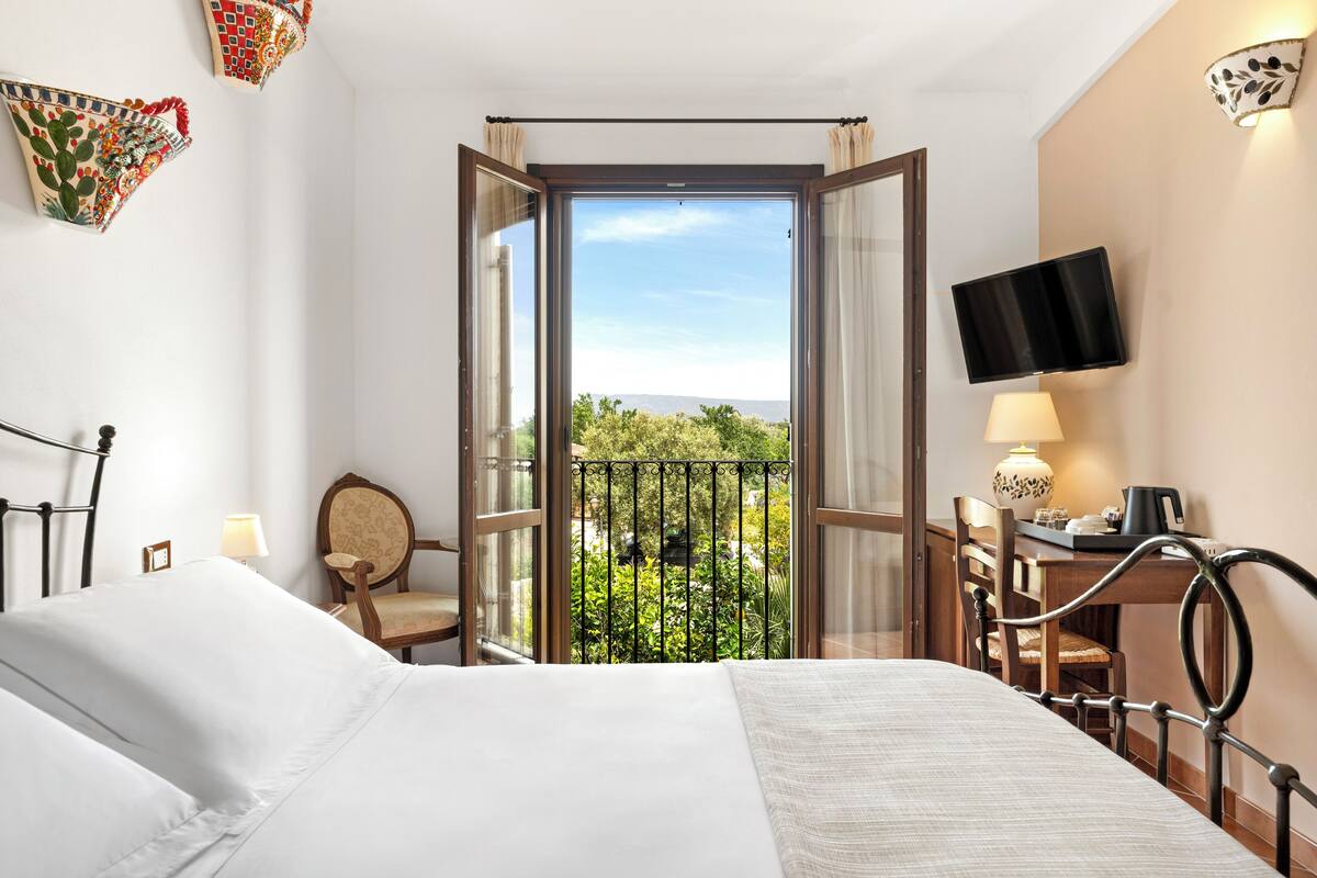 A hotel room with a bed, a desk with a chair, a TV, and a balcony with a view of greenery.