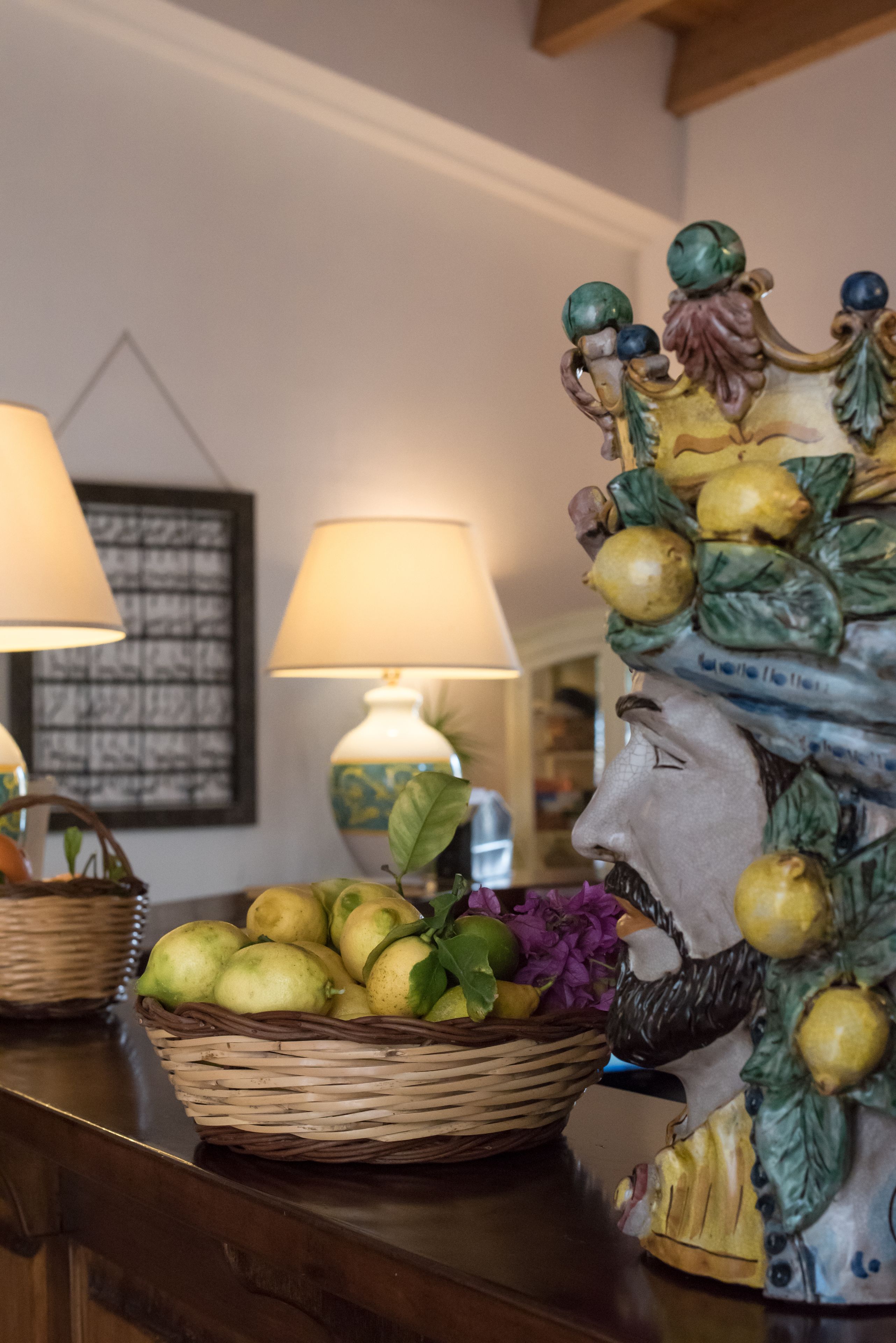 A room with a wicker basket filled with fruits and flowers on a wooden table, a decorative headpiece with fruits, and two lamps.