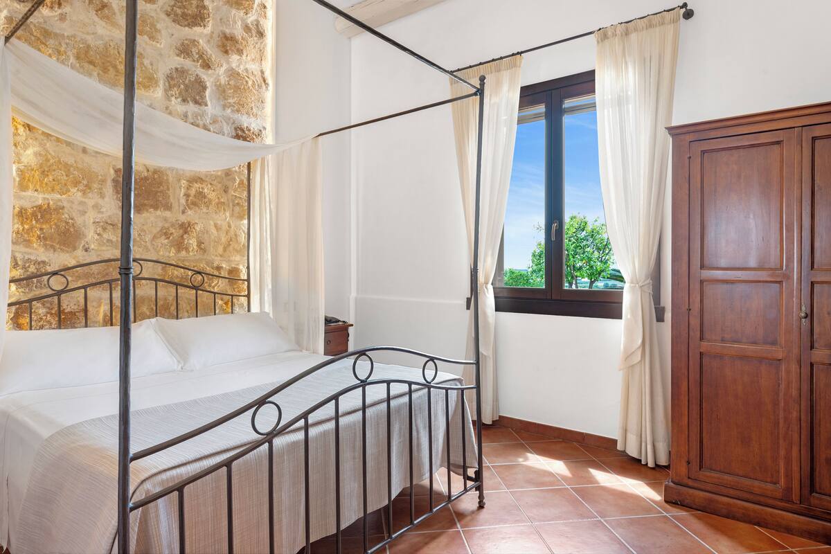 A four-poster bed with a stone wall backdrop, a wooden wardrobe, and a window with curtains.
