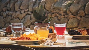 A table set with breakfast items and drinks, overlooking a coastal view with a stone wall and vegetation. - La Corte Del Sole (Noto)