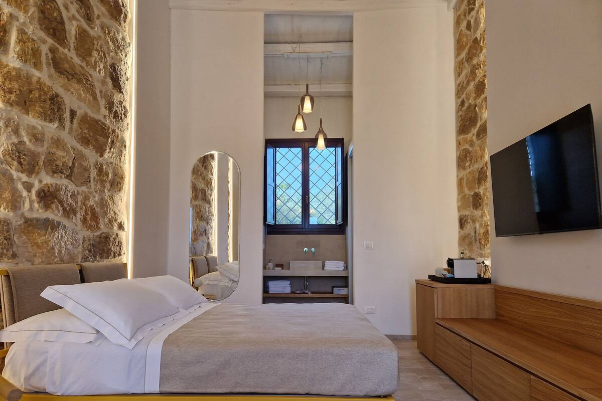 A bedroom with a stone wall, a bed with white linens, a wooden bedside table, a mirror, a window with a view, and a TV on the wall.