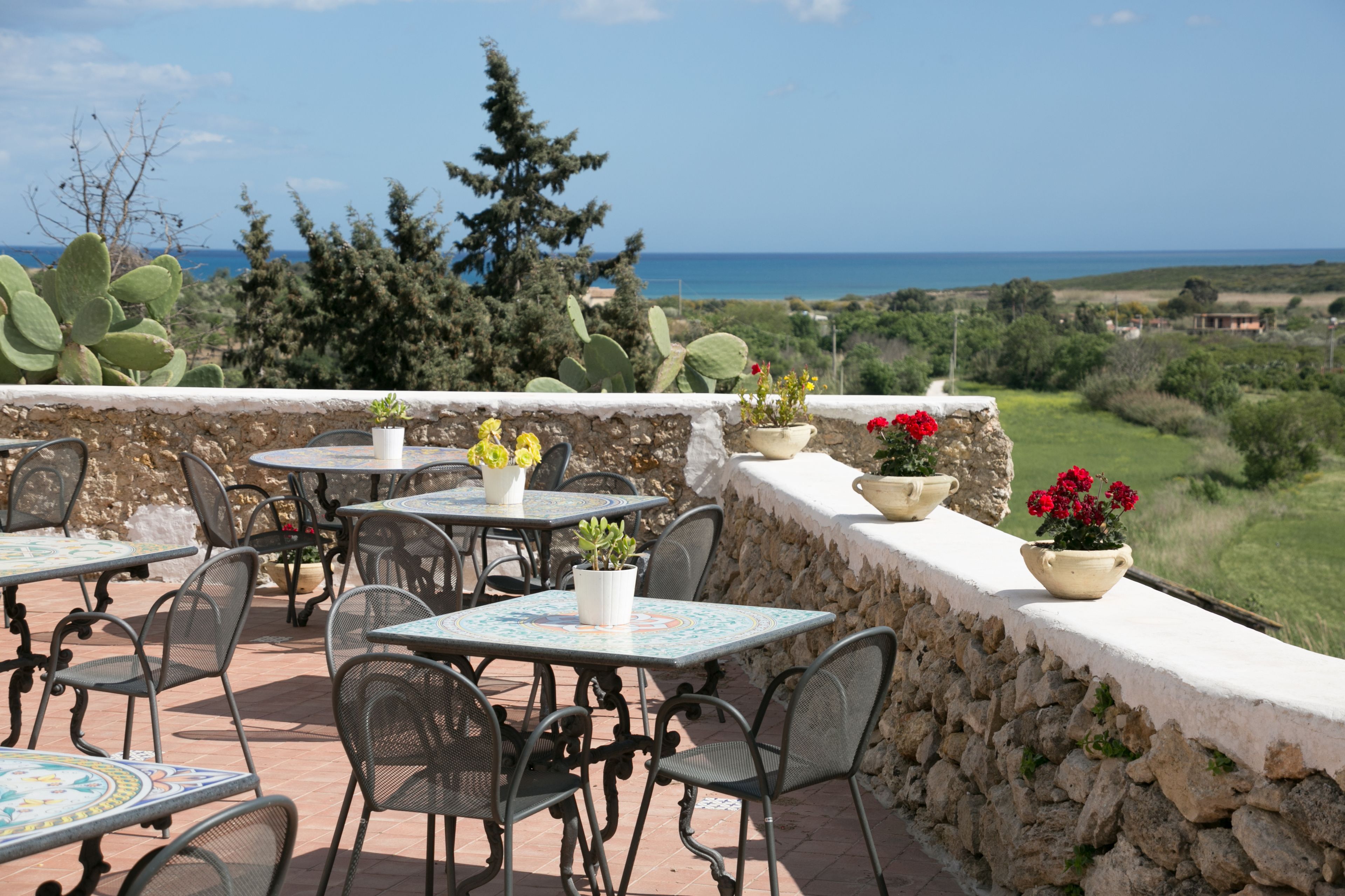 An outdoor patio with tables and chairs, potted plants, and a view of the sea.