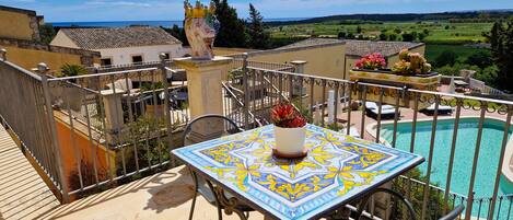 A balcony with a tiled table, chairs, and a view of a swimming pool and landscape.