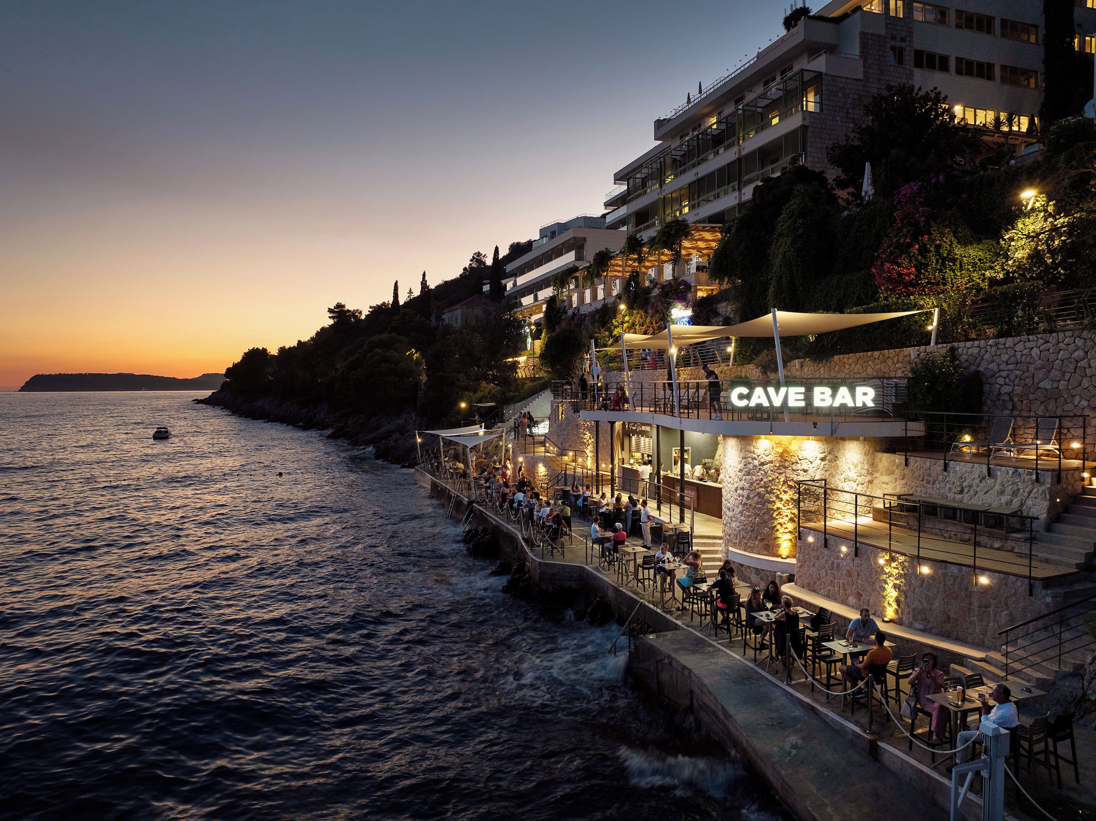 Eine Bar am Wasser mit Außenbereich und Meerblick, eine Fassade aus Stein und ein Schild mit der Aufschrift 'CAVE BAR'.
