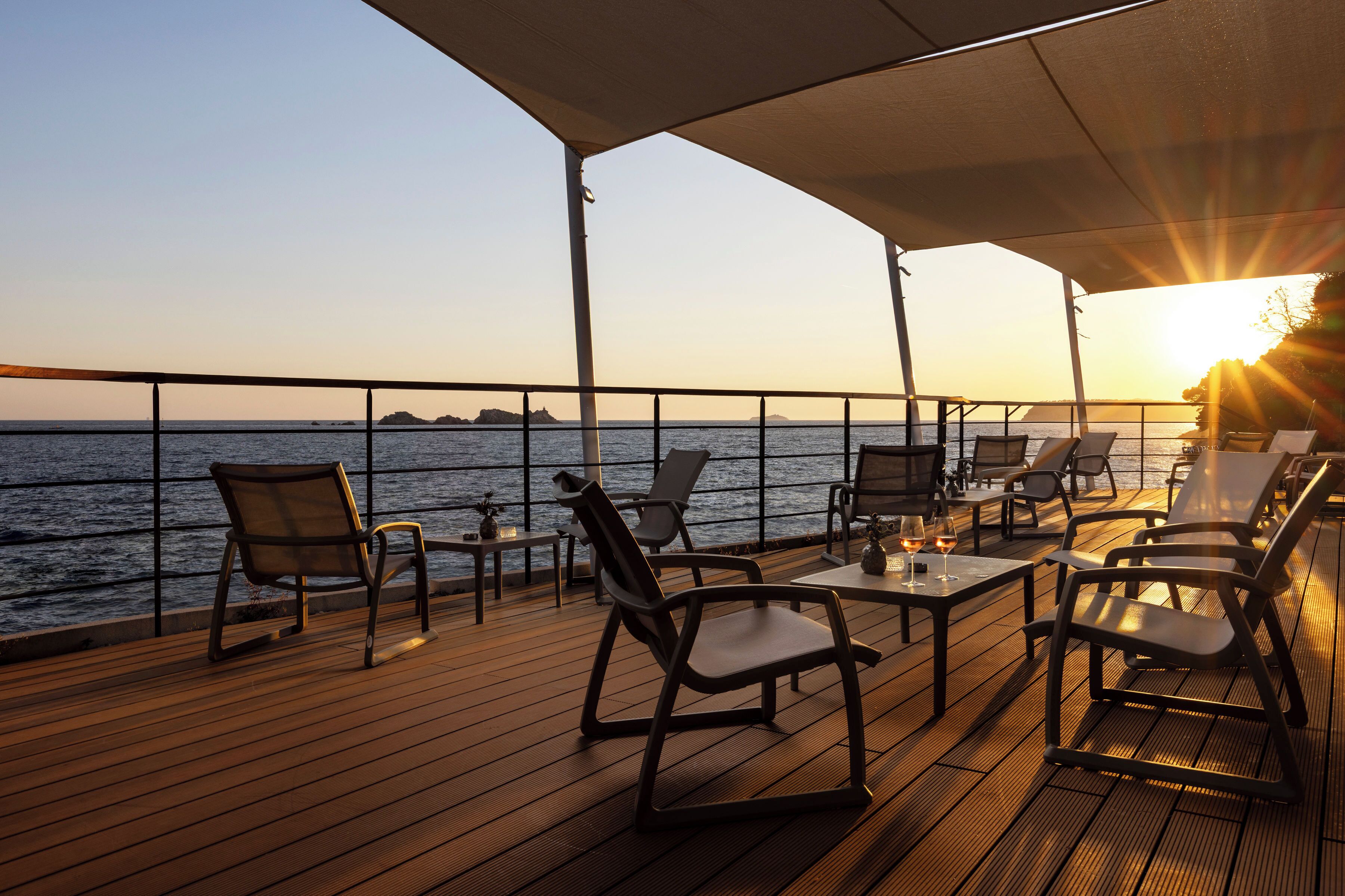 A deck with chairs and tables, overlooking a body of water with a sunset.
