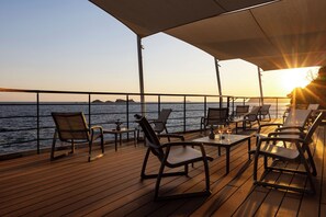 Een terras met stoelen en tafels, met uitzicht op een waterpartij in de zonsondergang.