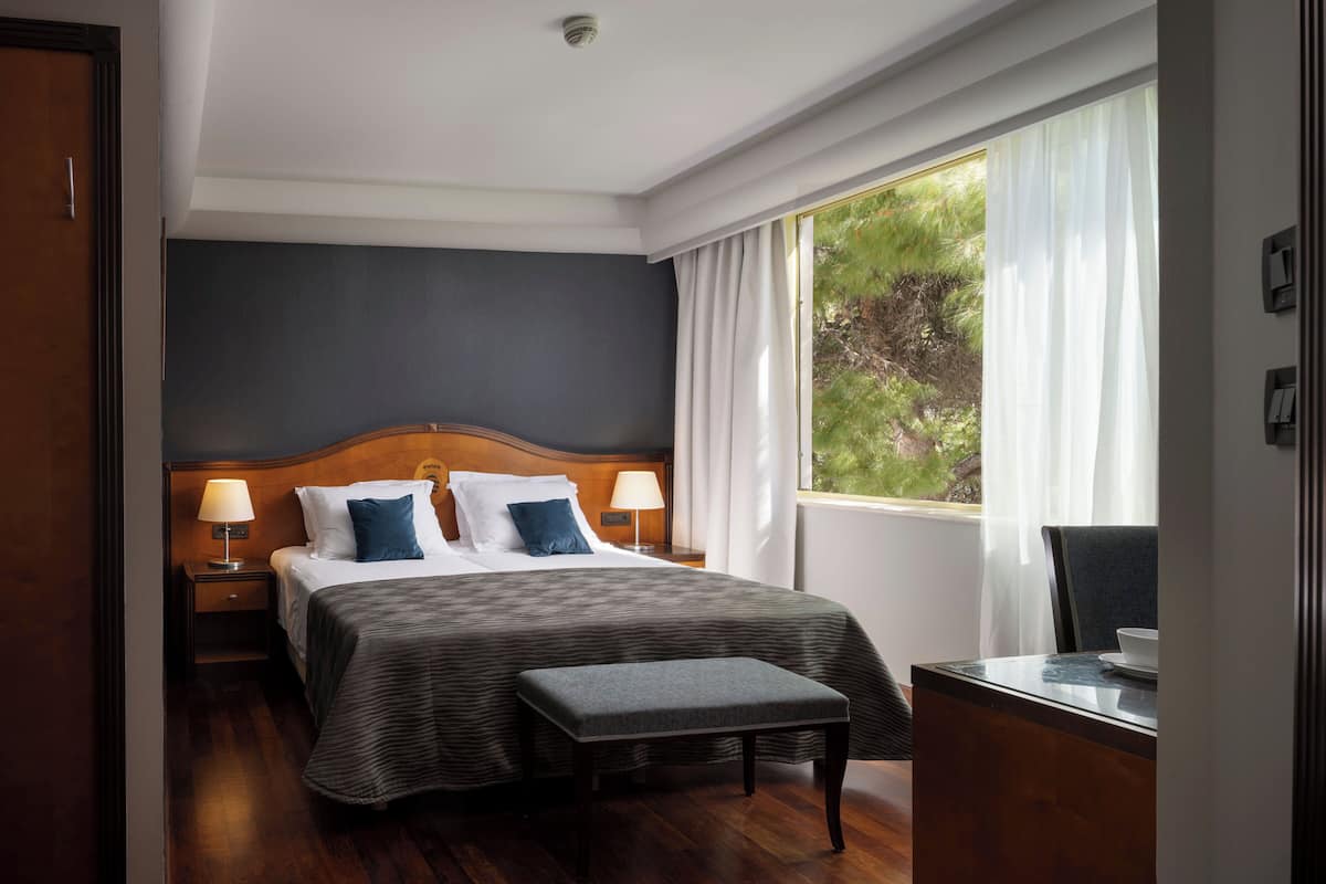 Quarto de hotel com uma cama grande, duas mesas de cabeceira, uma escrivaninha com cadeira e uma janela com cortinas.
