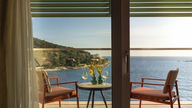A balcony with two chairs and a table, overlooking a coastal landscape with buildings and the sea.