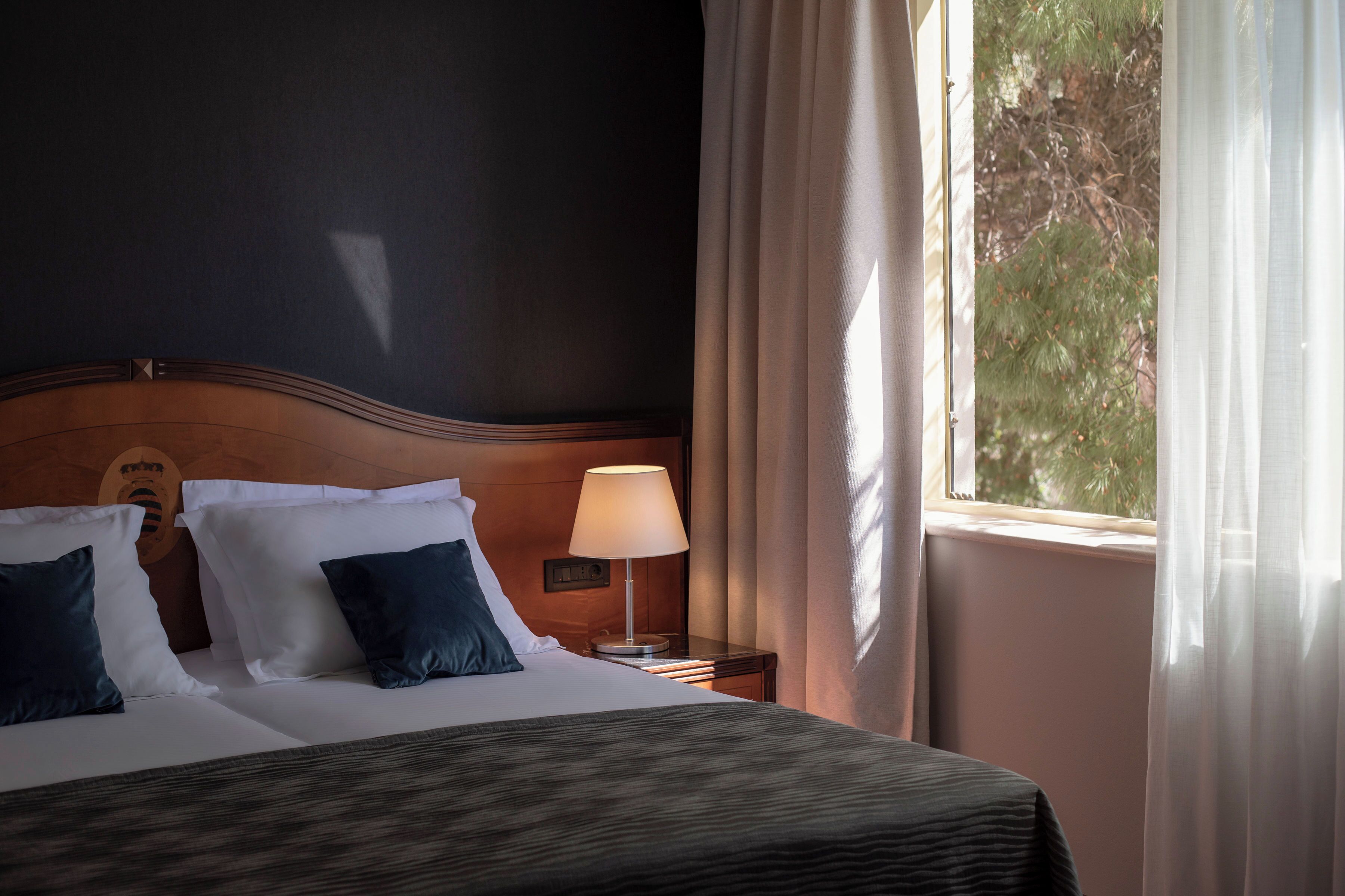 Una camera d'albergo con un letto, cuscini, un comodino con una lampada e una finestra con tende.