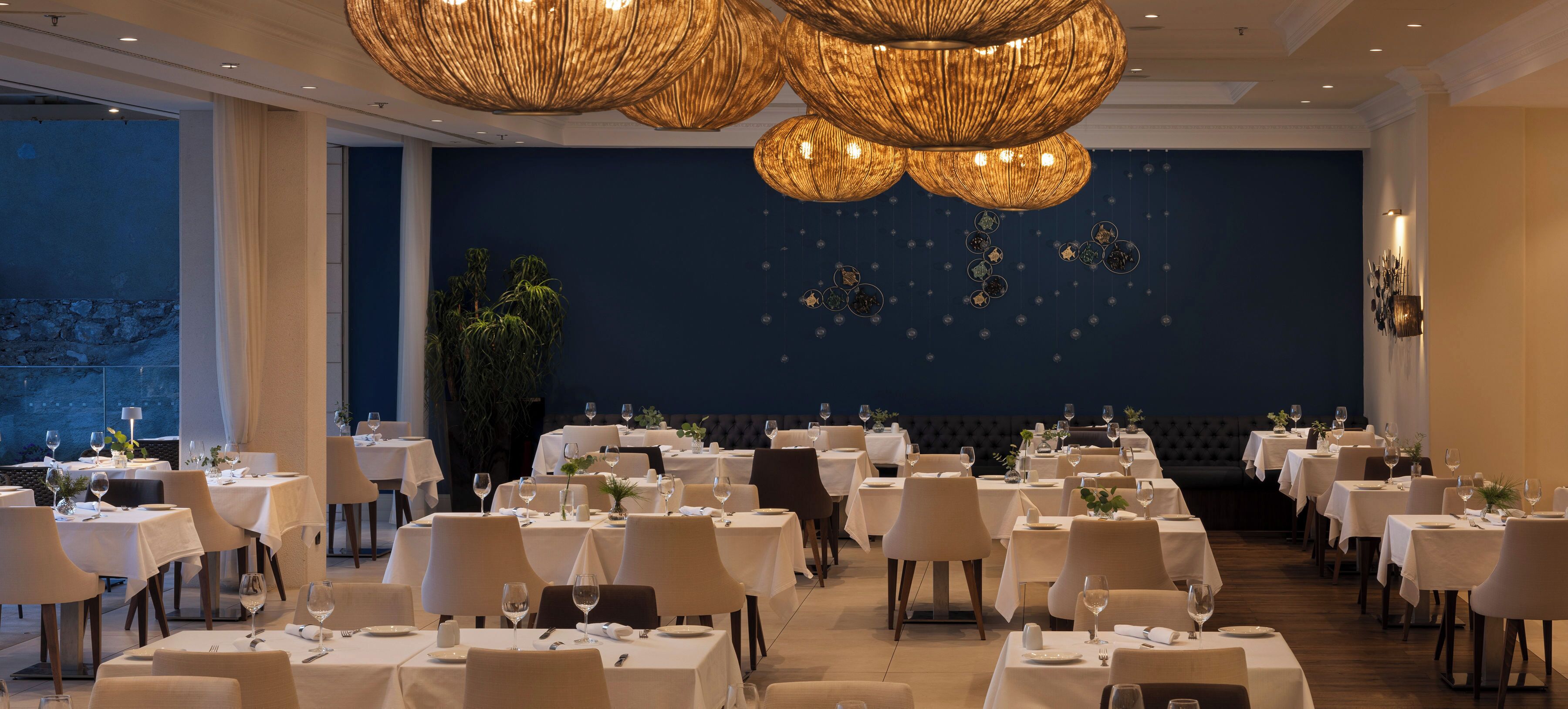 A restaurant with multiple tables set for dining, a large chandelier, and a dark blue wall with hanging decorations.