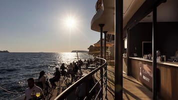 Ein sonnendurchfluteter Essbereich im Freien mit Tischen und Stühlen, mit Blick aufs Meer, sowie eine Bar mit dem Schild 'cave ba more'.