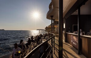 A sunlit outdoor dining area with tables and chairs, overlooking the sea, and a bar with a sign reading 'cave ba more'. - Hotel More (Dubrovnik)