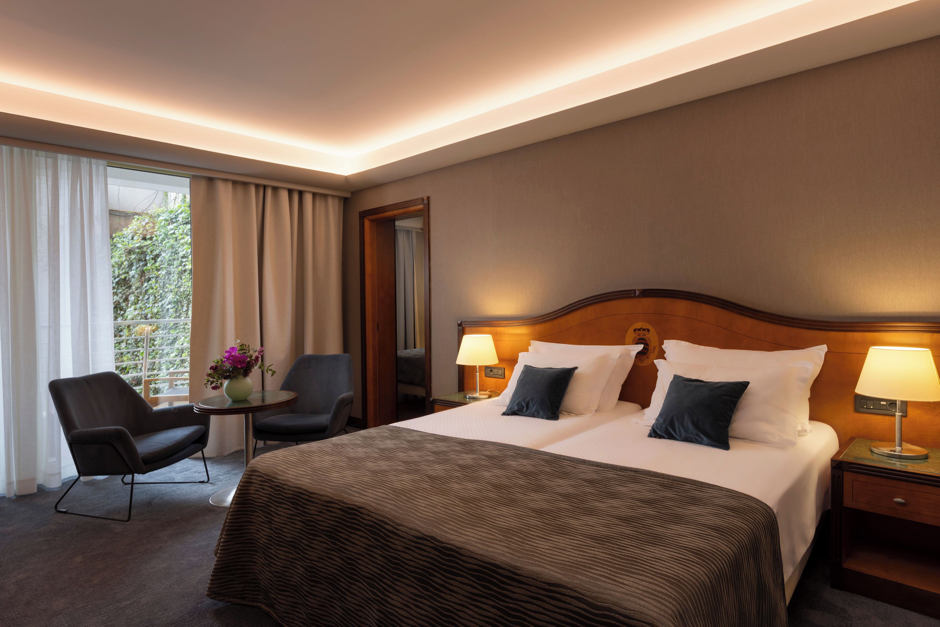Habitación de hotel con una cama doble, dos lámparas de noche, una silla, una mesita con un florero y vistas a zonas verdes por la ventana.