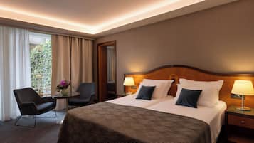 Habitación de hotel con una cama doble, dos lámparas de noche, una silla, una mesita con un florero y vistas a zonas verdes por la ventana.
