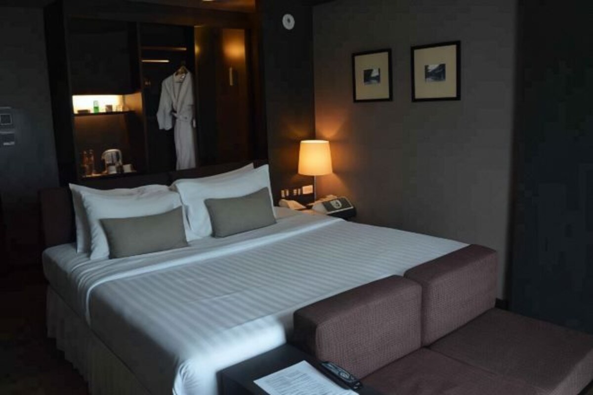 Habitación de hotel con cama, mesita de noche con lámpara, bata colgada en un armario y dos cuadros en la pared.