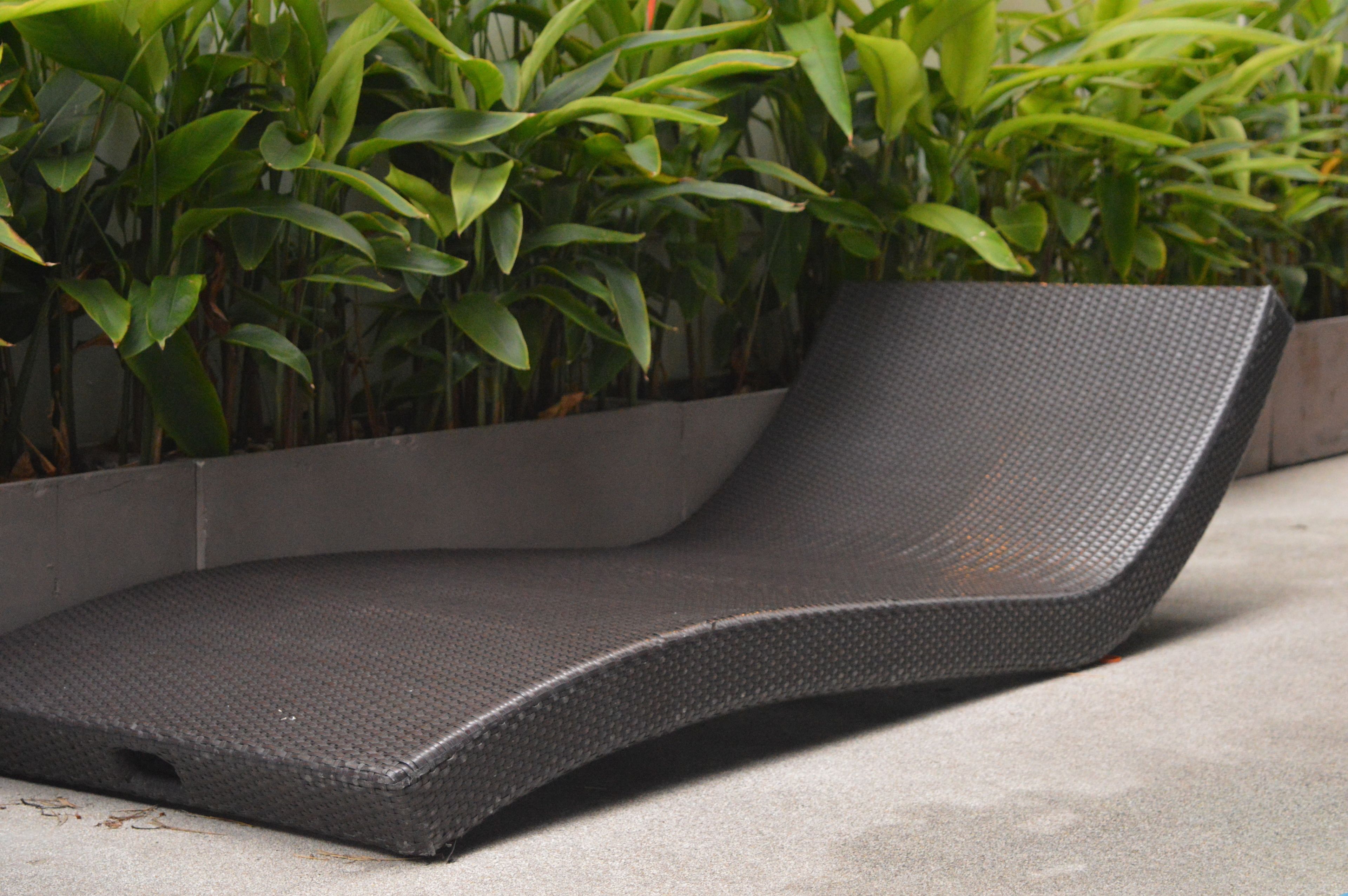 A modern lounge chair with a textured surface, positioned outdoors with a concrete floor and a planter with green foliage in the background.