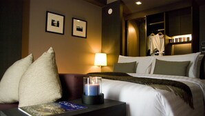 Habitación de hotel con una cama, almohadas, una mesita de noche con un libro y una vela, y un cuadro colgado en la pared.