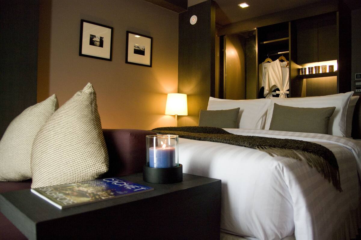 A hotel room with a bed, pillows, a bedside table with a book and a candle, and a wall-mounted picture.