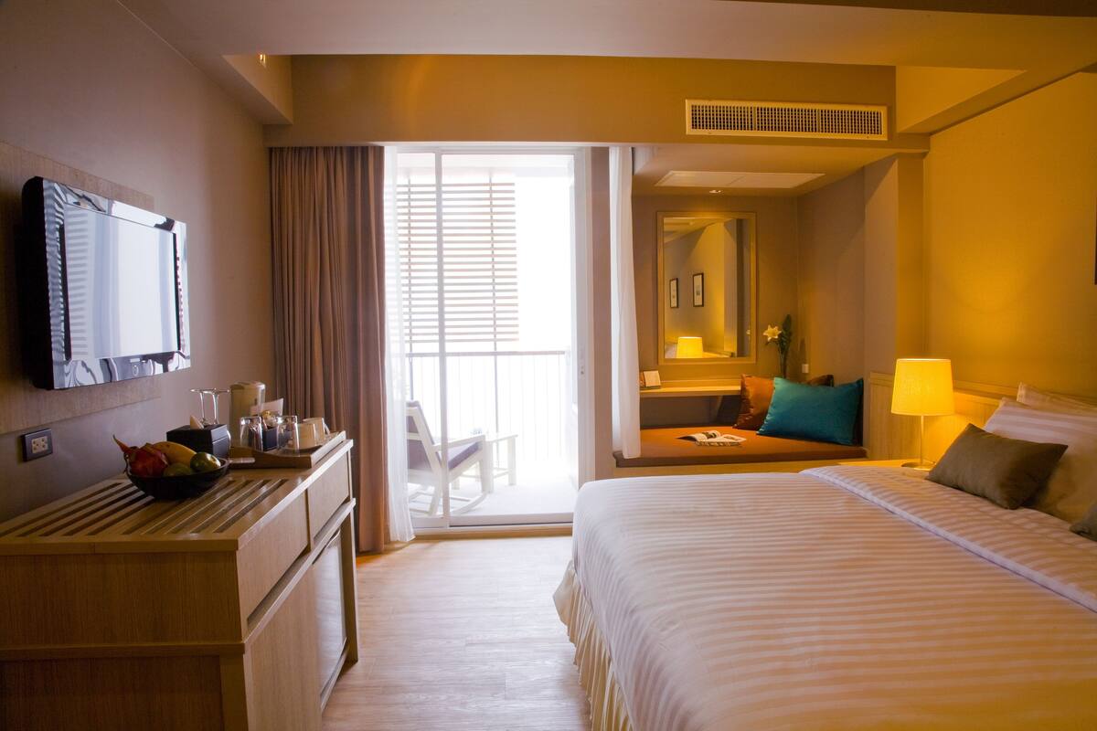 A hotel room with a bed, a desk with a bowl of fruit, a desk lamp, a chair, and a sliding door to a balcony.