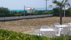 A beachside area with white plastic chairs and tables, a volleyball net, and a small white building.