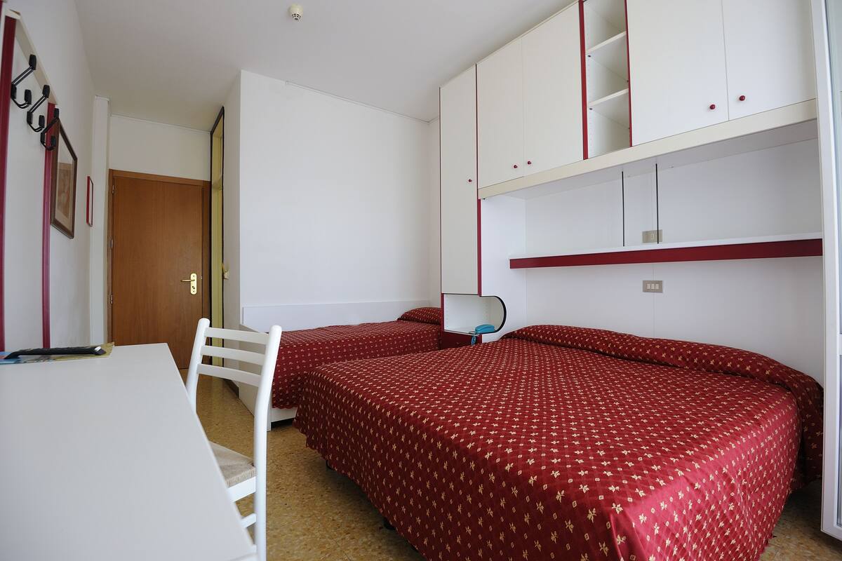 Ein Zimmer mit zwei Einzelbetten, einem Schreibtisch und einem Stuhl. Ein Kleiderschrank mit roten Akzenten und eine Tür sind im Hintergrund zu sehen.