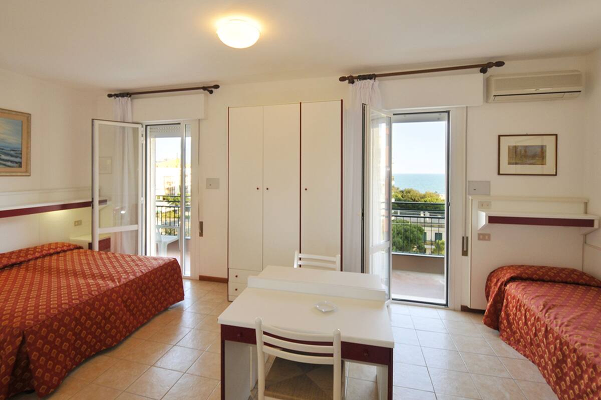 Ein Hotelzimmer mit zwei Betten, einem Tisch und einem Stuhl. Es gibt einen Balkon mit Blick auf das Meer.