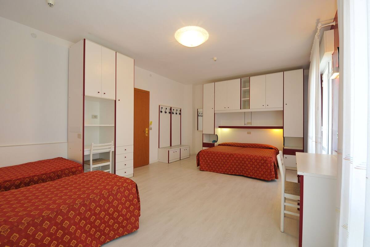 Ein Zimmer mit zwei Einzelbetten, einem Schreibtisch und einem Stuhl. Es gibt einen Schrank mit Spiegel und eine Tür mit Schloss.