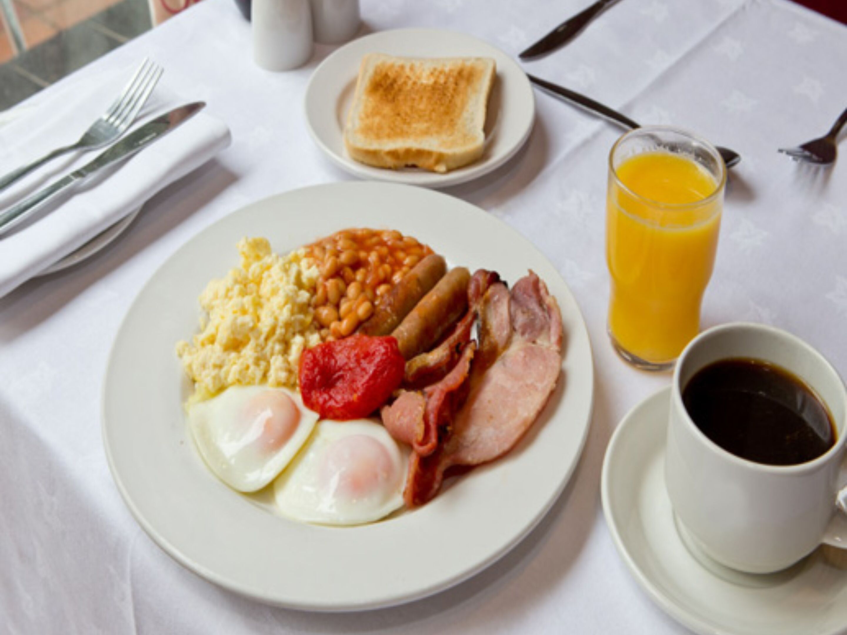 Un plato de desayuno con huevos, frijoles, tomates, salchichas y tocino, acompañado de tostadas, jugo de naranja y café.
