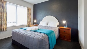 A neatly made bed with a blue blanket and white pillows, two bedside tables with lamps, a window with curtains, and a dark textured wall. - Aotea Motel (Christchurch)