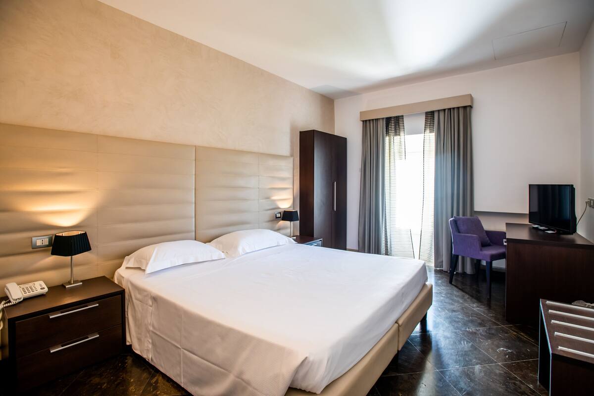 Une chambre d’hôtel avec un grand lit, un bureau avec une chaise, une télévision et une fenêtre avec des rideaux.