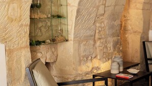 A stone wall interior with a glass shelf, a cushioned chair, a table with plates and a book, and a small lamp.