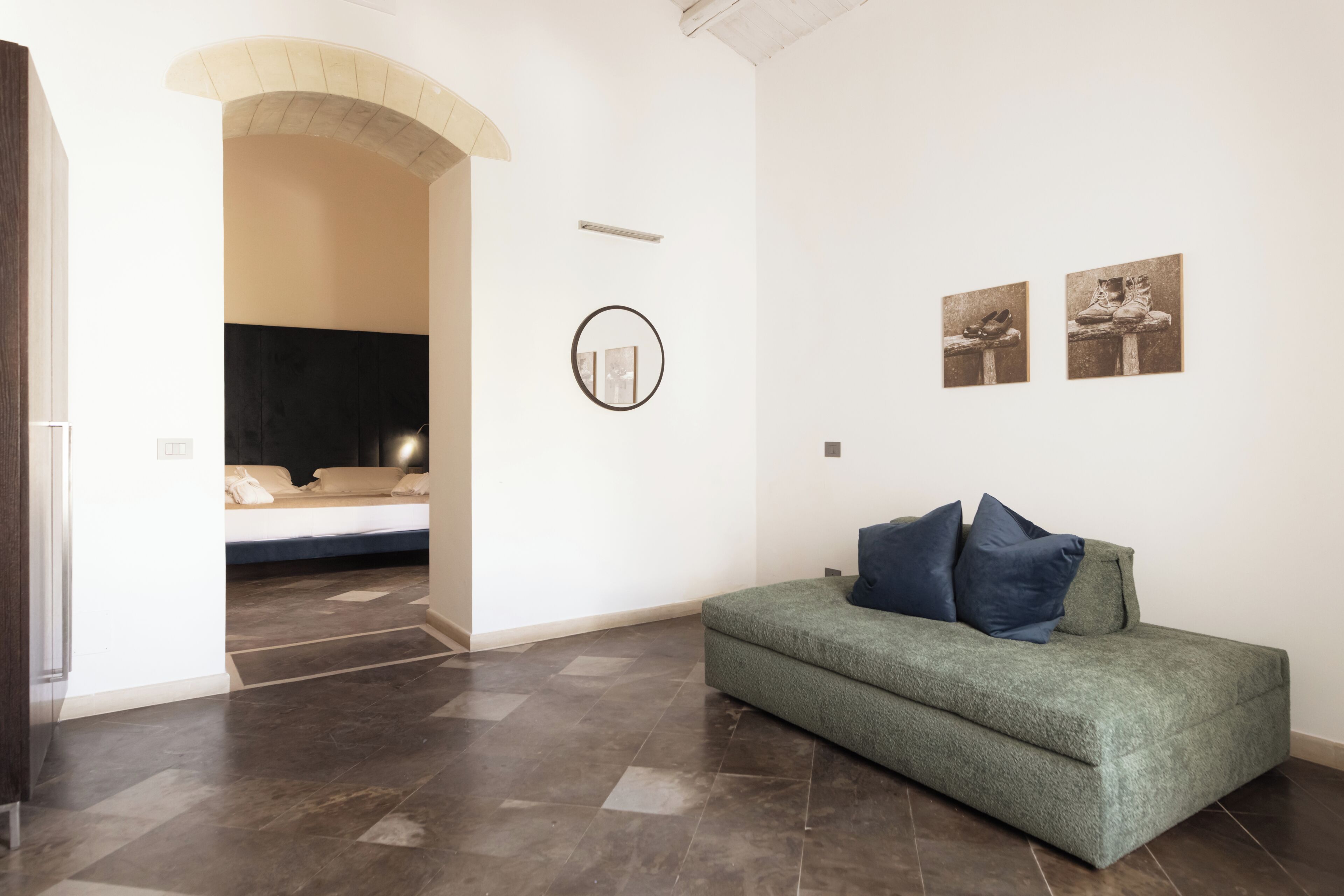 Quarto com sofá verde, cama com cabeceira, espelho circular e duas fotos emolduradas na parede.