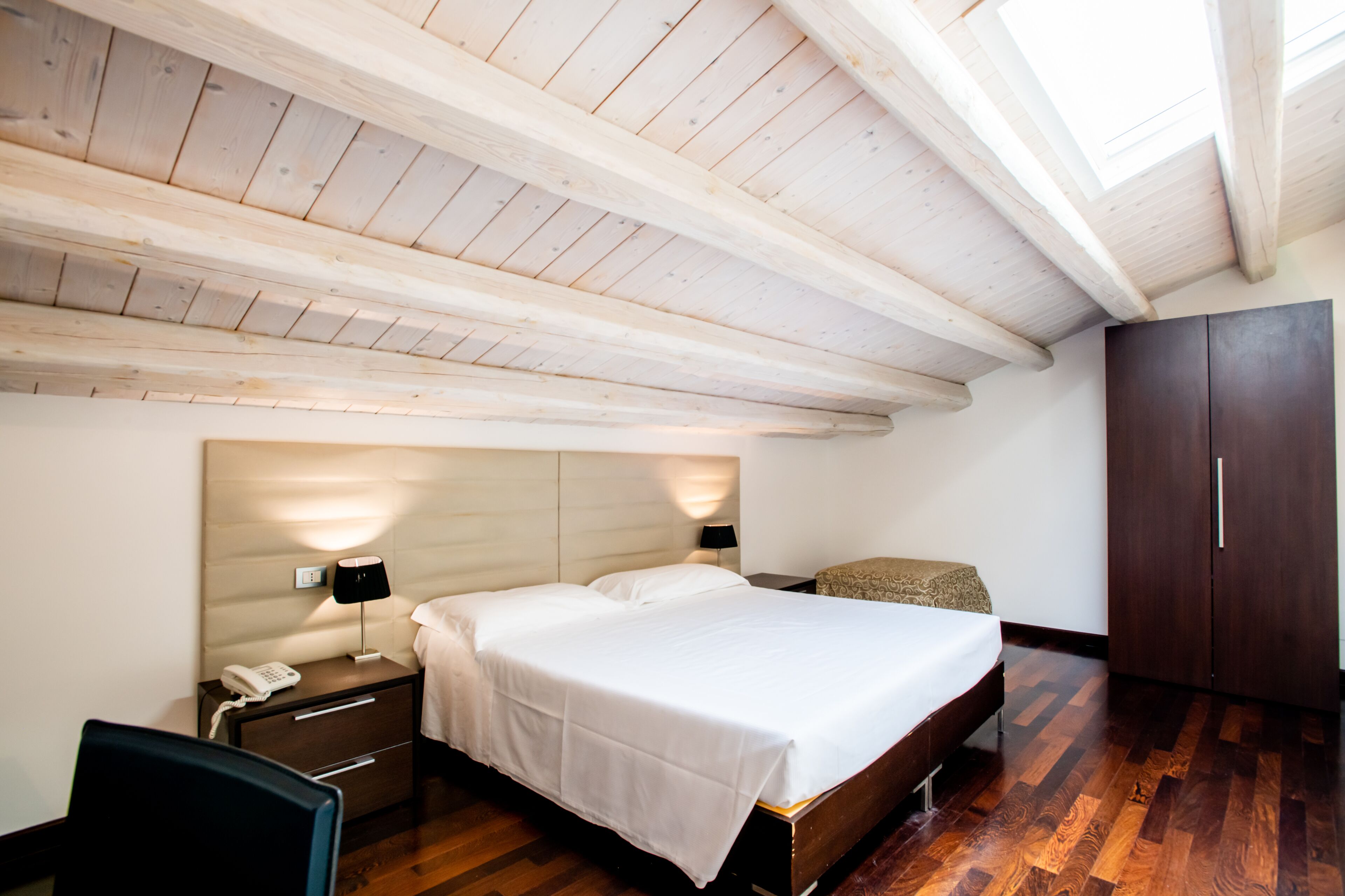 Quarto com teto de madeira, cama com roupa de cama branca, mesa de cabeceira com abajur e guarda-roupa.