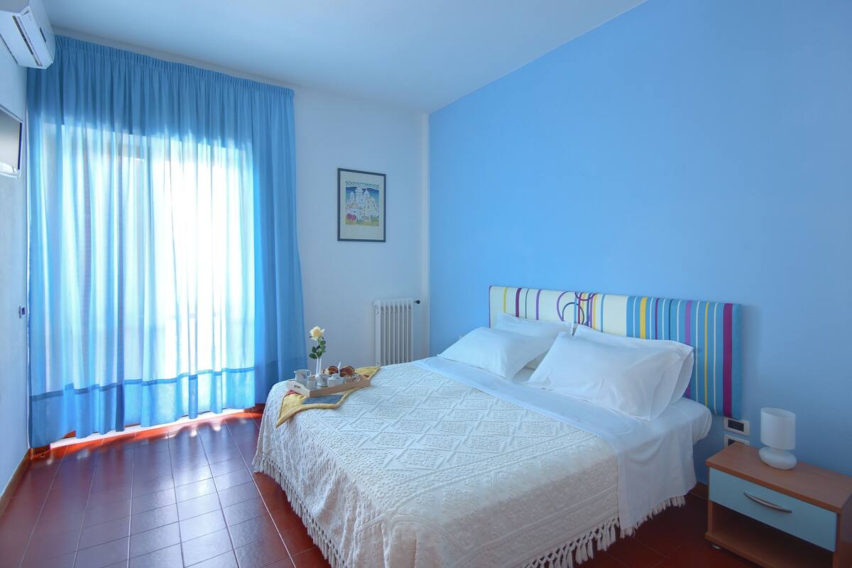 A bedroom with a bed, a bedside table, a nightstand, a window with curtains, a radiator, and a framed picture on the wall.