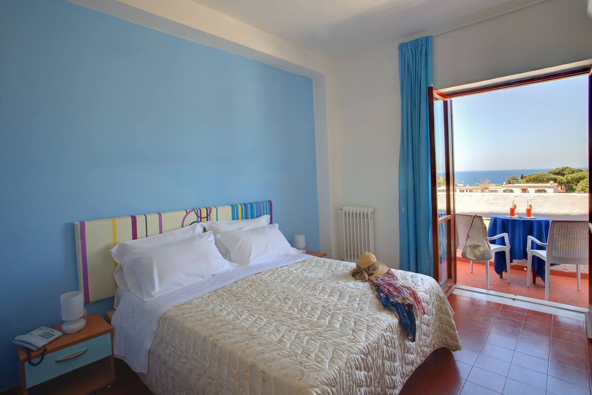 A bedroom with a bed, a nightstand, a window with a view of the beach, and a table with chairs outside.