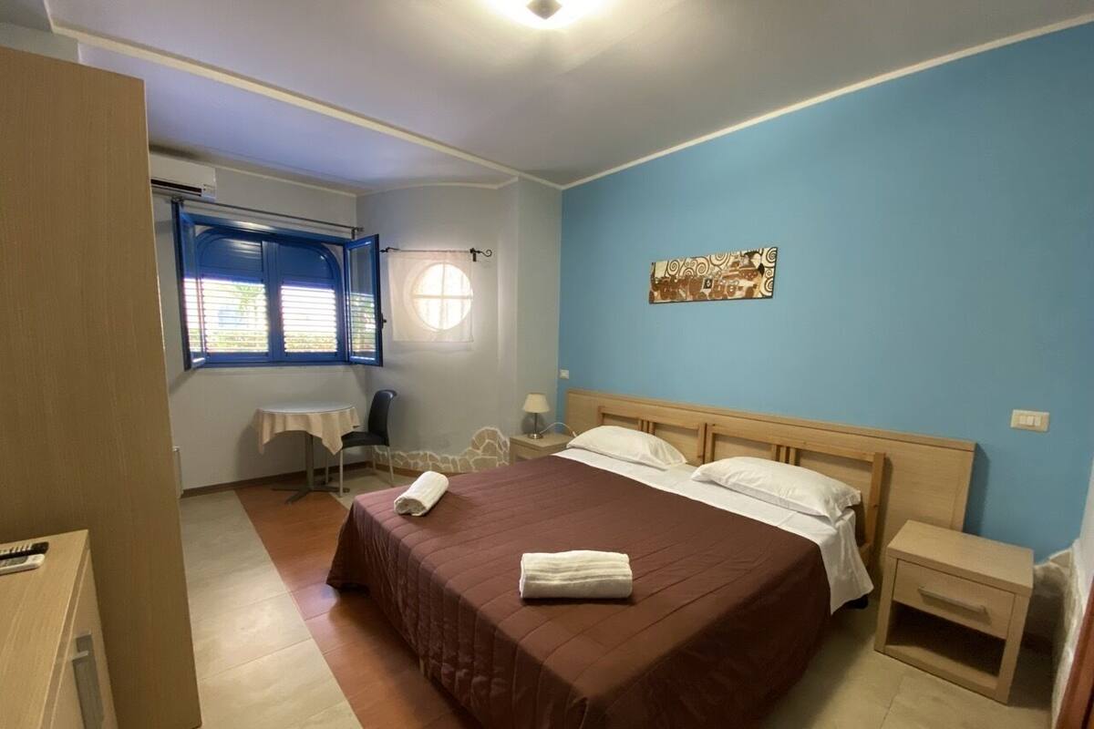 Een slaapkamer met een groot bed, twee nachtkastjes, een klein tafeltje, een raam met zonwering en een aan de muur bevestigd kunstwerk.