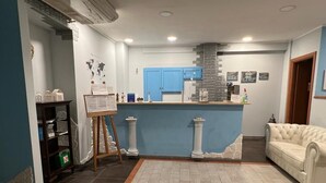 A reception area with a blue counter, a white sofa, and a small display stand. - Kassiopea Aparthotel (Giardini Naxos)